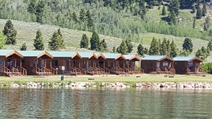 Una hilera de cabañas de madera con un muelle que se adentra en un lago, rodeado por una ladera boscosa.