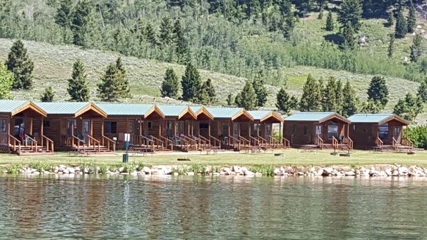 A row of wooden cabins with a dock extending into a lake, surrounded by a forested hillside. - Elk Haven Camping Cabin 11 (West Yellowstone)