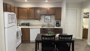 Una cocina moderna con armarios de madera, un refrigerador blanco, un microondas y un lavavajillas. Hay una mesa de comedor con dos sillas y una pequeña planta en maceta.