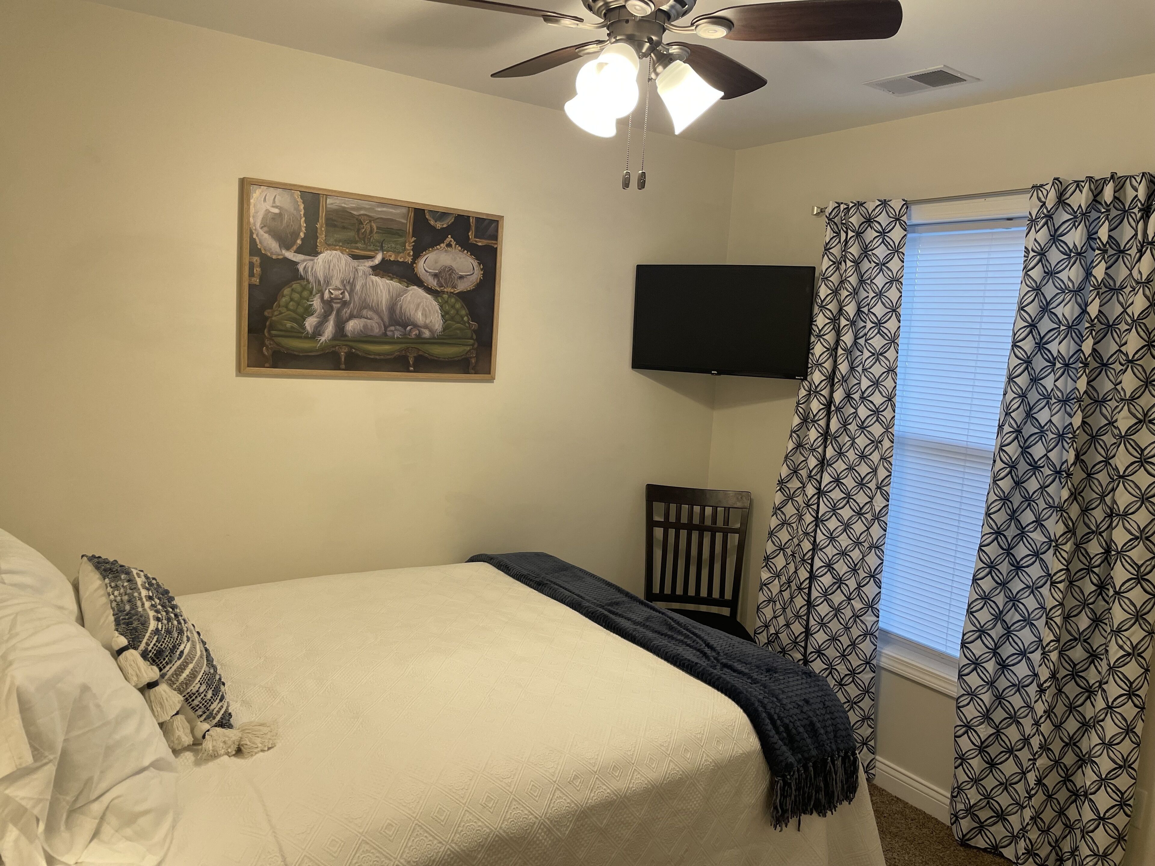 A bedroom with a bed, a ceiling fan, a TV mounted on the wall, and patterned curtains.