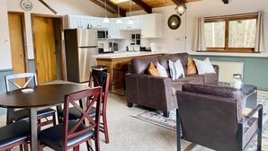 Un salon chaleureux avec un canapé en cuir marron, une table à manger avec des chaises, et un coin cuisine doté de meubles blancs et d’un réfrigérateur en acier inoxydable.
