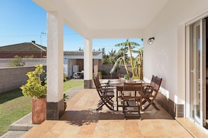 A covered patio with a dining table and chairs, a potted plant, and a view of a house and palm trees.