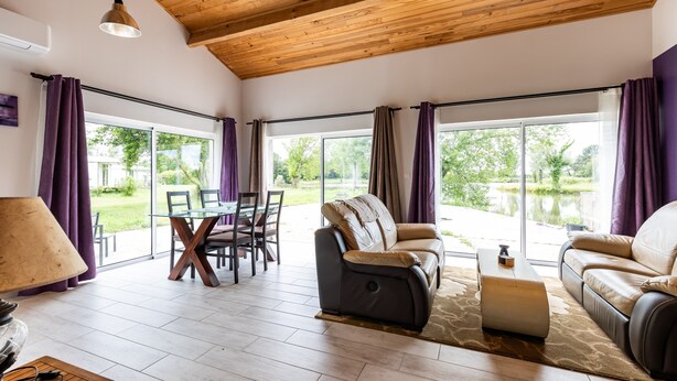 Ein großzügiger Wohnbereich mit Holzboden, großen Fenstern mit violetten Vorhängen, einem Ledersofa, einem Esstisch mit Stühlen und einer Lampe.
