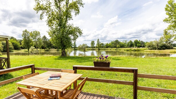 Una terraza de madera con una mesa y sillas, con vistas a un lago rodeado de árboles y un cielo nublado.