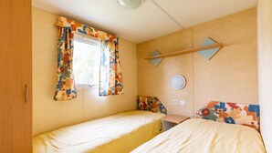 Una cama individual con edredón amarillo, una ventana con cortinas de colores, un espejo fijado a la pared y una mesita con una silla.