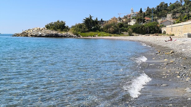 Una playa con agua cristalina, un litoral rocoso y edificios al fondo.