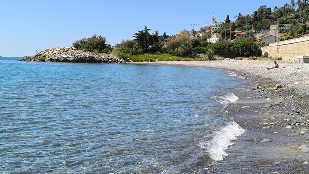 Ein Strand mit klarem Wasser, felsigem Ufer und Gebäuden im Hintergrund.