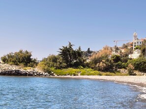 A coastal area with a rocky shoreline, dense greenery, and buildings in the background.