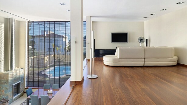 Una sala de estar moderna con piso de madera, un sofá blanco, un televisor de pantalla plana y un balcón con vistas a edificios y una piscina.