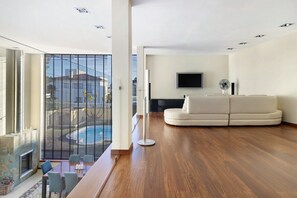 Uma sala de estar moderna com piso de madeira, um sofá branco, uma TV de tela plana e uma varanda com vista para edifícios e uma piscina.