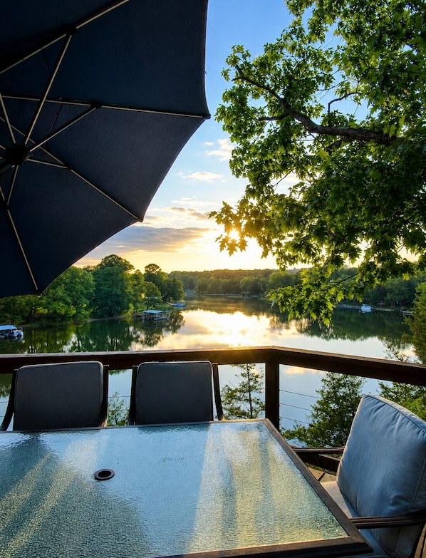 A table with chairs and an umbrella overlooking a lake with trees and a sunset.