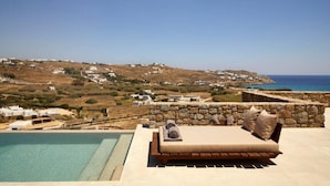 Un salon de bord de piscine avec un mur en pierre et un lit, offrant une vue sur un paysage côtier avec des maisons et la mer.