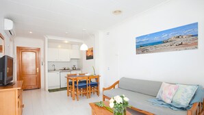 Una sala de estar con un sofá, una mesa de centro con flores, una zona de comedor con sillas y una mesa, y una cocina al fondo.
