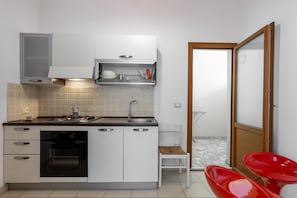 Une cuisine compacte avec des meubles blancs, un four encastré, un évier et un petit coin repas agrémenté d’une chaise rouge.