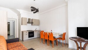 Une cuisine moderne avec des meubles blancs, une table à manger entourée de chaises orange, un téléviseur posé sur un meuble en bois et un canapé orange à motifs.