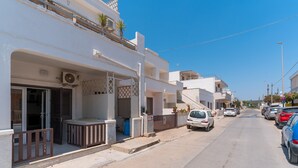 Une rue résidentielle bordée d’immeubles blancs, avec des voitures garées et un ciel bleu dégagé.