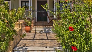 A stone pathway leading to a building with a white pergola, potted plants, and a red flower.
