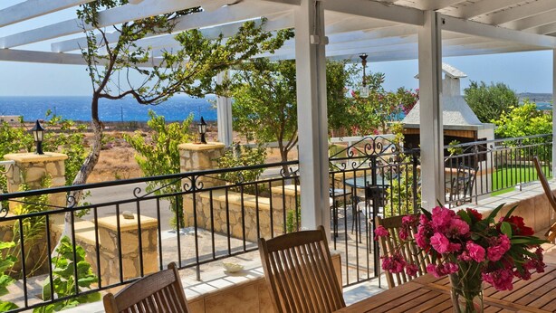 A covered outdoor patio with a wooden table and chairs, a hanging light fixture, and a view of the ocean.