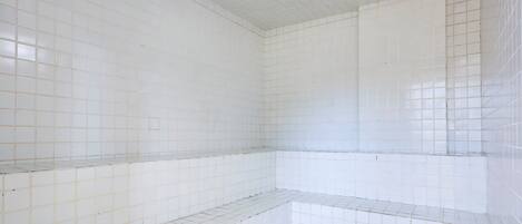 Una habitación con paredes y piso de baldosas blancas, que presenta una esquina con estante bajo.