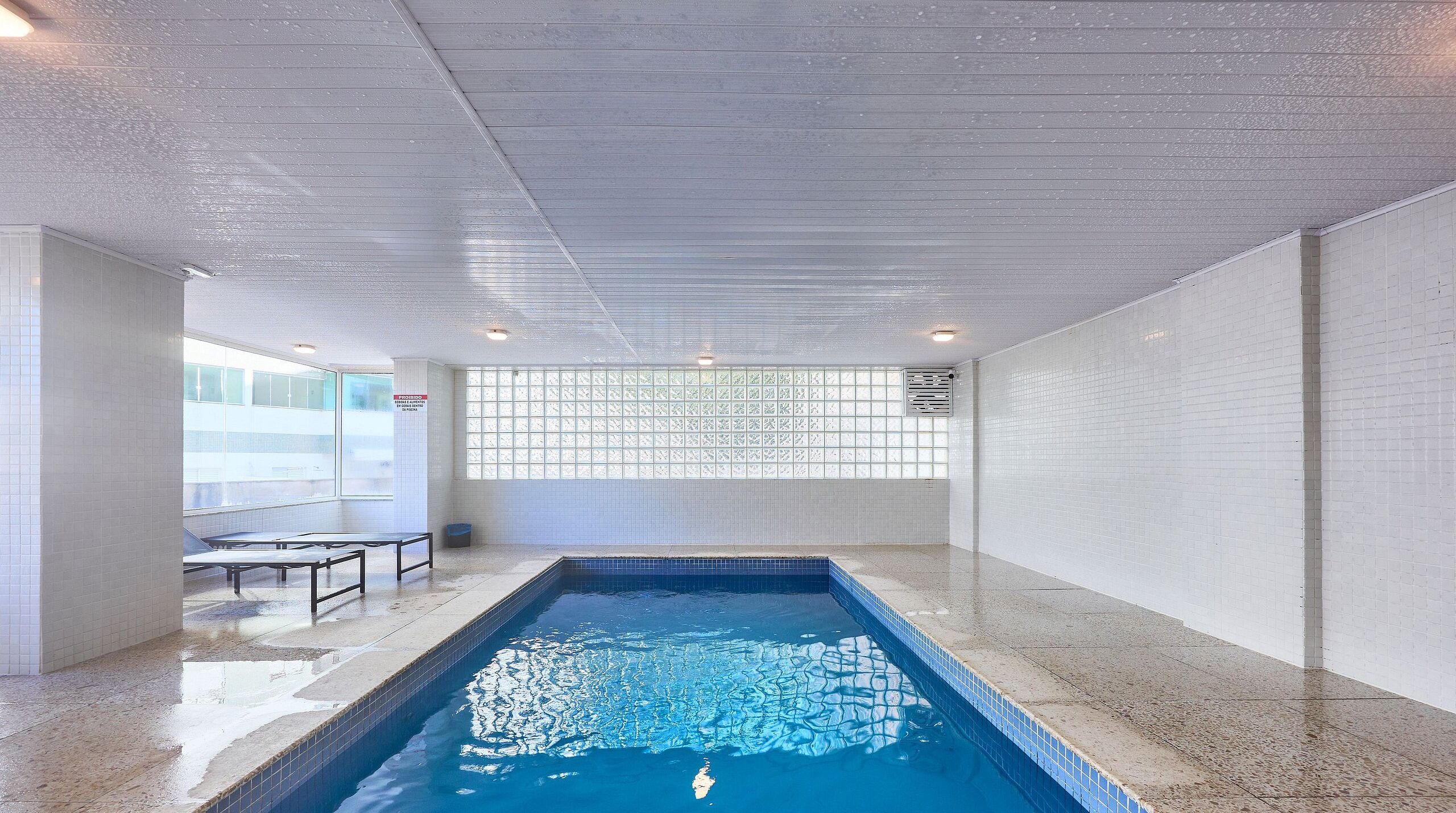 An indoor swimming pool with a tiled floor, a bench, and a window with a grid pattern.