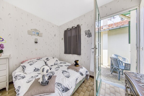 Un dormitorio con cama, mesita de noche, cómoda, ventana con cortinas y vistas a un espacio exterior con sillas.