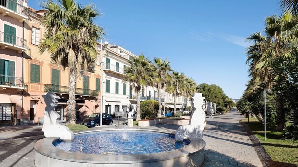 A public square with a fountain, palm trees, and buildings with balconies. - Apartment 'Smart Home Loano' with Wi-Fi and Air Conditioning (Loano)
