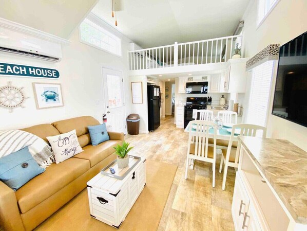 A spacious living area with a sofa, coffee table, and a dining table with chairs. There is a kitchen in the background with white cabinets and a black refrigerator.