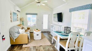 A spacious living room with a high ceiling, a ceiling fan, a large window, a television, a dining table with chairs, and a sofa with a throw blanket. - Vacation Cottage North Myrtle Beach #68 (North Myrtle Beach)