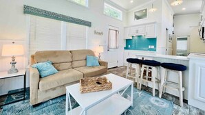 A spacious living room with a beige sofa, a coffee table, and a kitchen area with white cabinets and a teal backsplash. - Vacation Cottage North Myrtle Beach #65 (North Myrtle Beach)