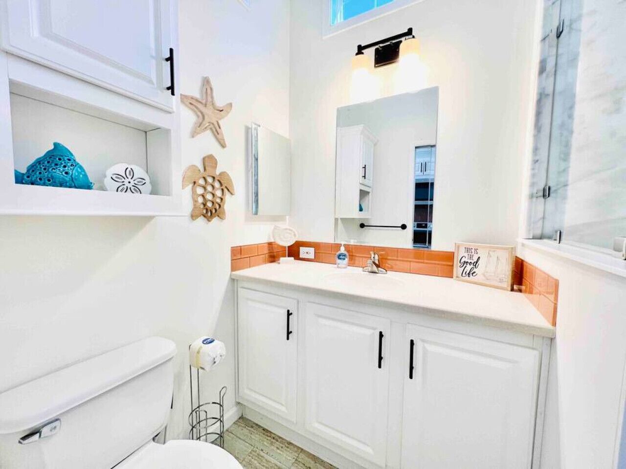 Ein Badezimmer mit Toilette und Waschbecken in Weiß. Das Waschbecken hat eine orangefarbene Arbeitsplatte, darüber hängt ein Spiegel. An der Wand befinden sich Dekorationen und ein Handtuchspender.