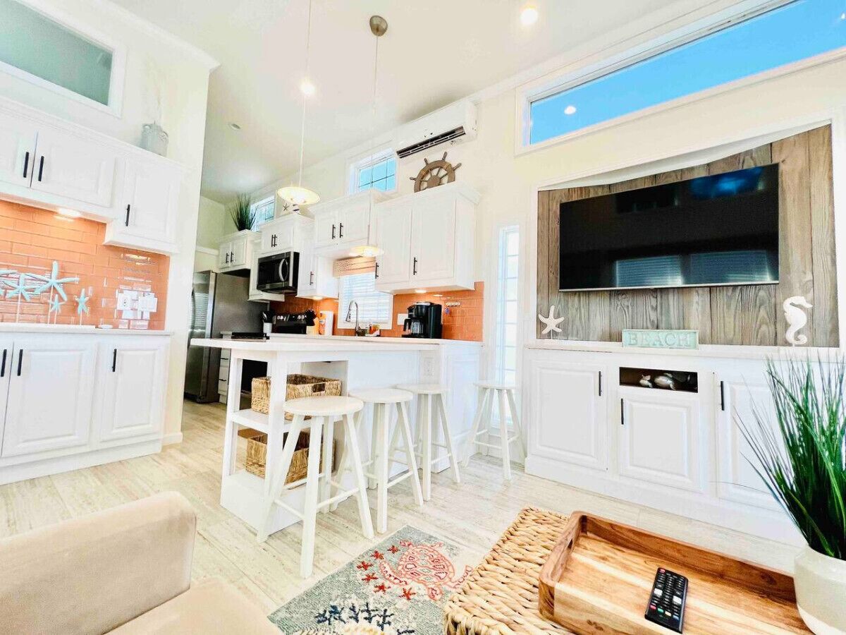 Una cocina moderna con armarios blancos, una isla central con taburetes, un televisor grande y una pared de acento de madera.
