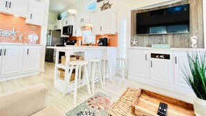 A modern kitchen with white cabinets, a central island with stools, a large TV, and a wooden accent wall. - Vacation Cottage North Myrtle Beach #67 (North Myrtle Beach)