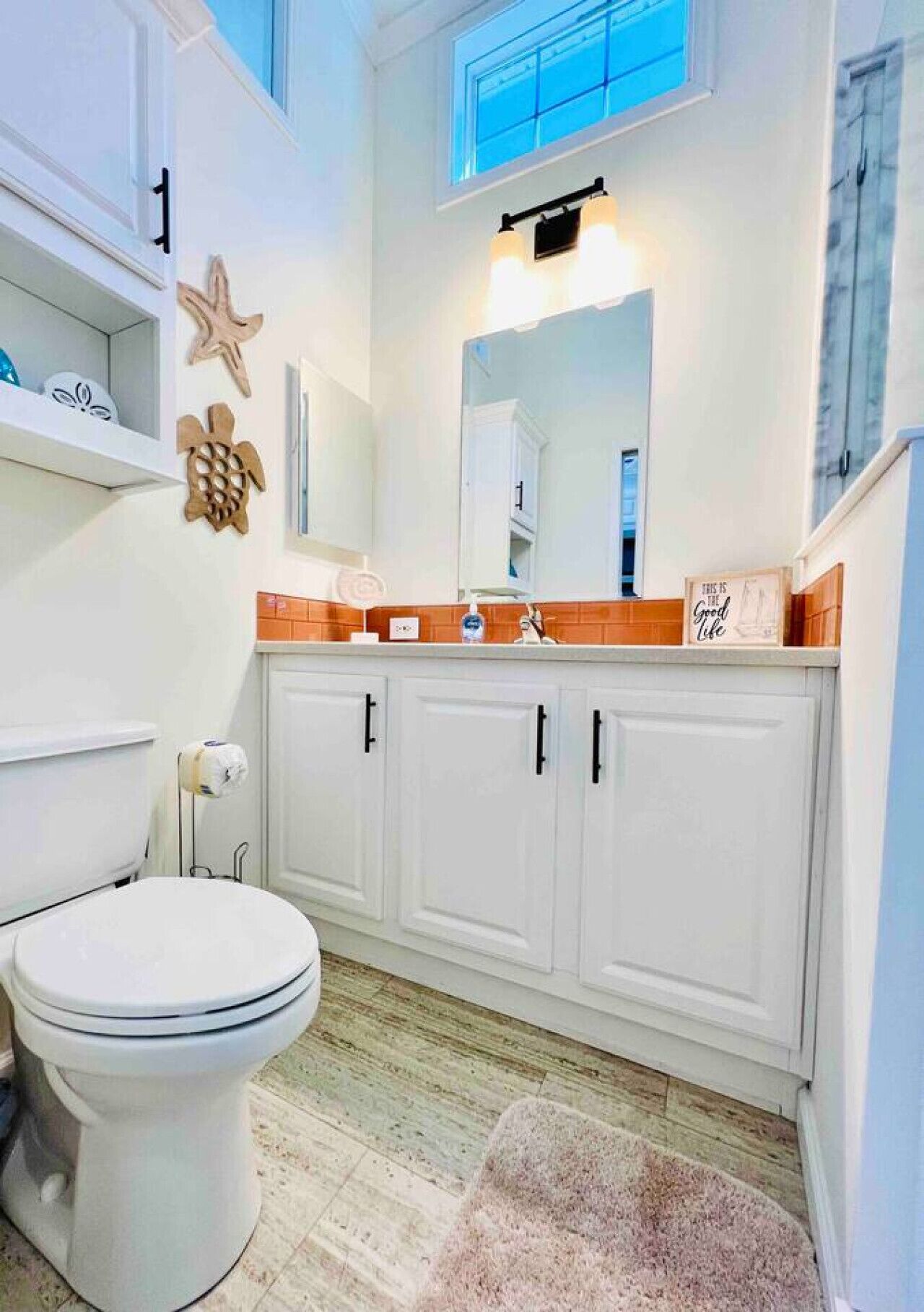 Une salle de bain avec des toilettes, des meubles blancs, un miroir et une décoration en forme d’étoile de mer en bois.