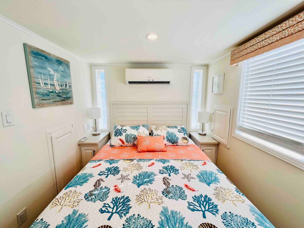 A bedroom with a bed, a nightstand, a wall-mounted air conditioner, a window with blinds, and a painting on the wall.