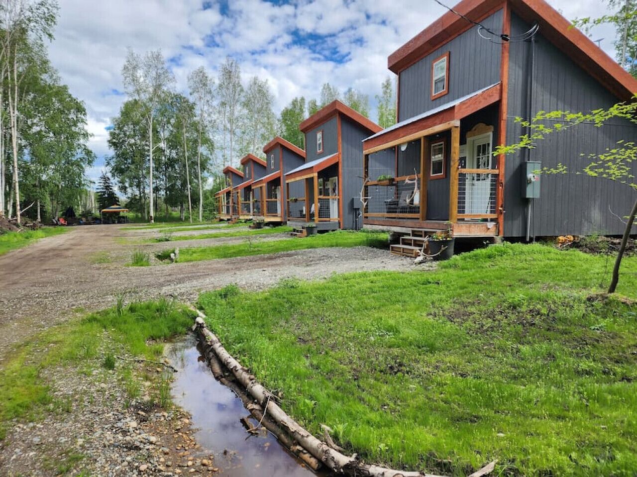 Une rangée de chalets modernes aux façades en bois et aux grandes fenêtres, situés dans un espace verdoyant, près d’un petit ruisseau.
