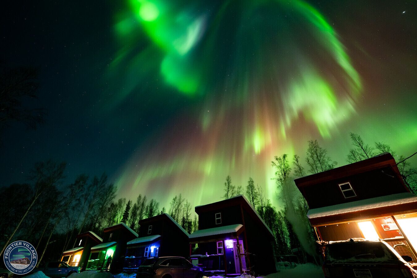 Una hilera de cabañas con nieve en el suelo, bajo un cielo nocturno con auroras verdes.
