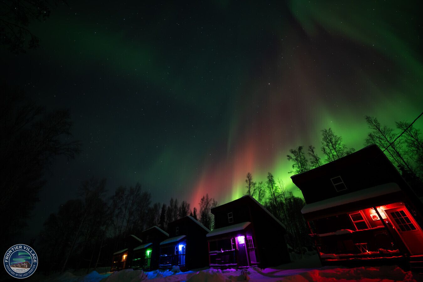 Une rangée de cabanes illuminées de lumières colorées, se détachant sur un ciel nocturne où scintillent les aurores boréales.