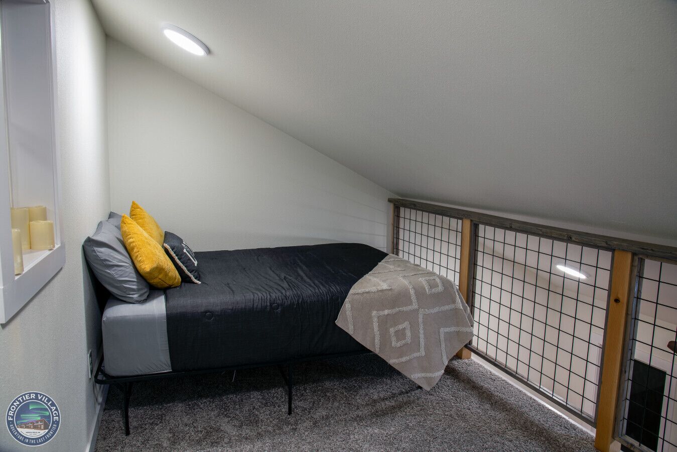 A small, single-bed room with a sloped ceiling, a window with candles, a grey carpet, and a decorative wall panel.