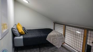 A small, single-bed room with a sloped ceiling, a window with candles, a grey carpet, and a decorative wall panel.