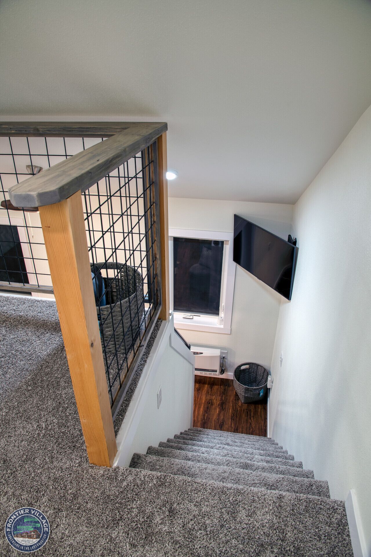Una escalera con barandilla de madera y pasamanos de metal, escalones alfombrados, una ventana con persianas, un televisor montado en la pared y una cesta en el suelo.