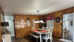 A dining area with a wooden paneled wall, a red tablecloth, white chairs, and a hanging light fixture. - Cozy Cottage with Pool Table! (Rocky Mount)