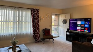 A living room with a large flat-screen TV displaying a streaming service interface, a window with blinds, a patterned curtain, a decorative wall piece, and a cozy seating area with a sofa and armchair. - Cozy Cottage with Pool Table! (Rocky Mount)