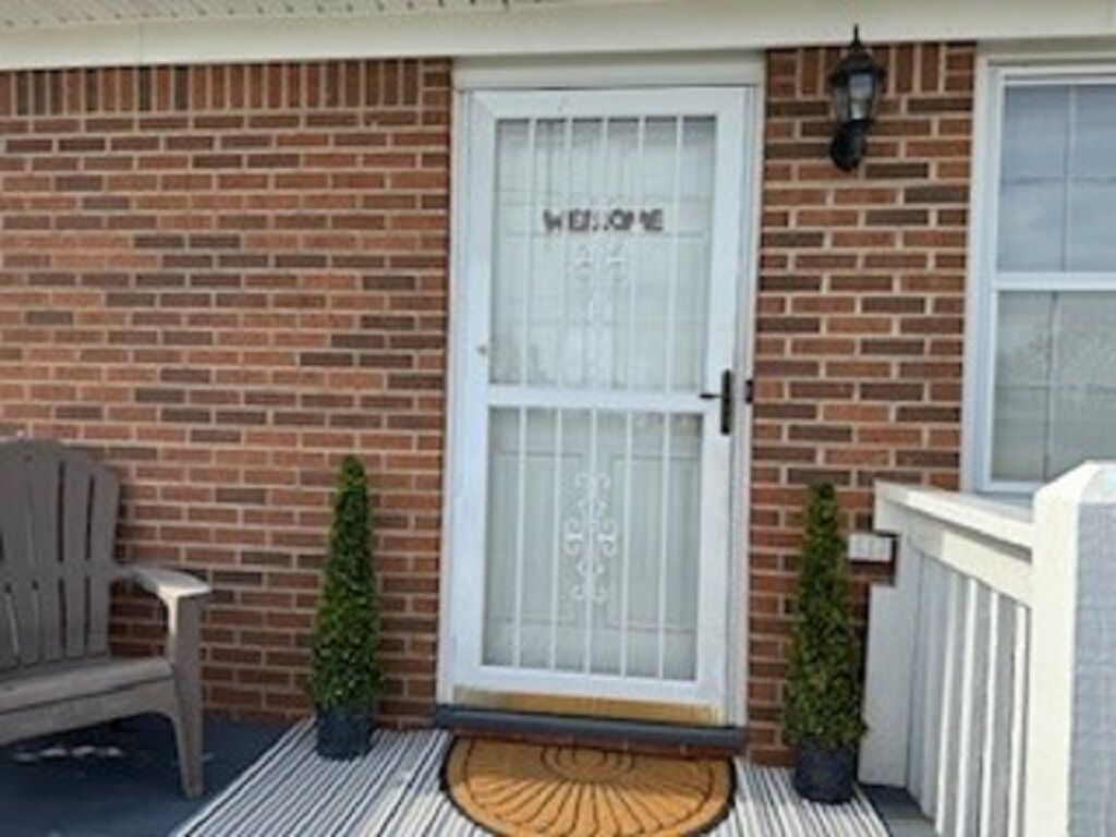 A brick house with a white door marked "WELCOME," a bench, two potted plants, and a white fence.