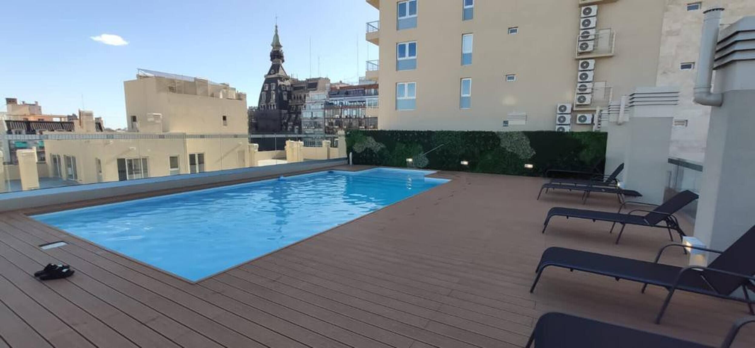 Una piscina en la azotea con sillones de descanso, una terraza de madera y vistas a la ciudad de fondo.