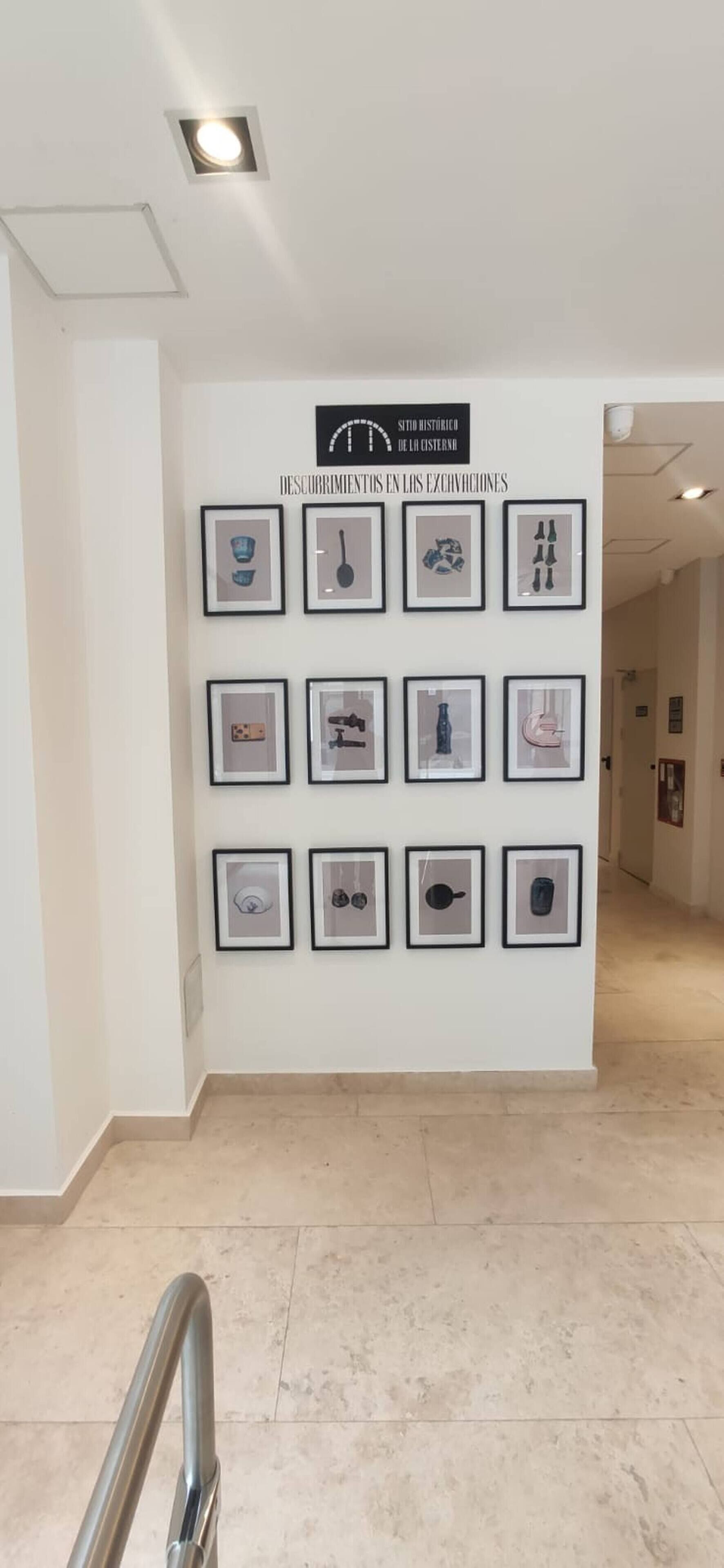 A hallway with a series of framed pictures on the wall, a sign above them, and a metal handrail in the foreground.