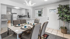 Een moderne keuken met witte kasten, een eethoek met een tafel voor vier personen en een grote potplant bij de deur.