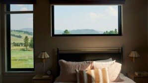 A bedroom with a large window offering a view of a green landscape and hills. There is a bed with multiple pillows and two bedside tables with lamps.