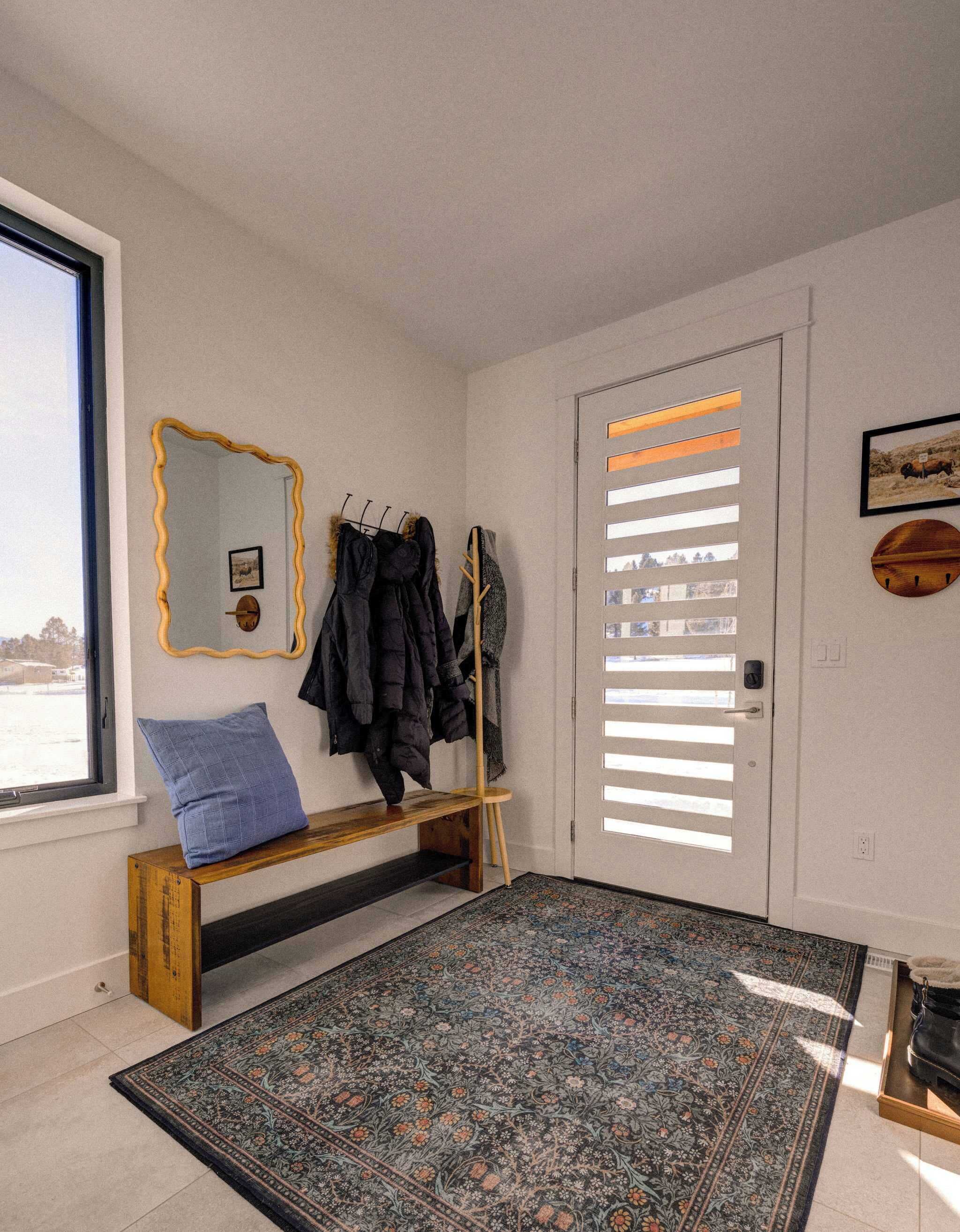 A room with a bench, a coat rack with jackets, a patterned rug, a mirror with a wavy frame, and a door with horizontal blinds.
