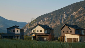 A modern two-story house with a garage, situated in a grassy field with mountains in the background. - Mountain Home By Aspects | Sauna Access + Firepit (Big Sky)