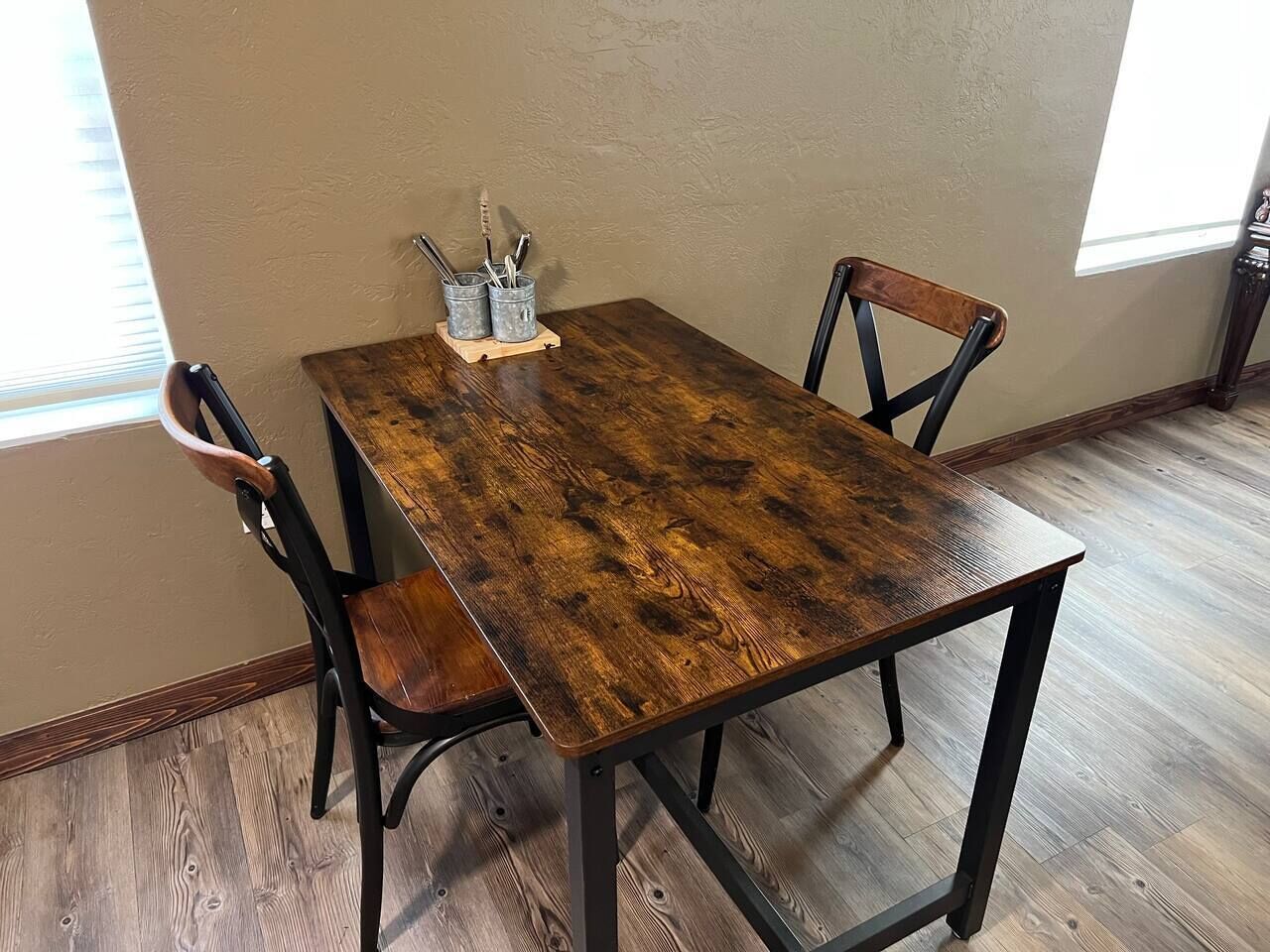 A rectangular wooden dining table with metal legs, accompanied by two chairs with wooden seats and metal frames. A small metal tray with utensils is placed on the table.